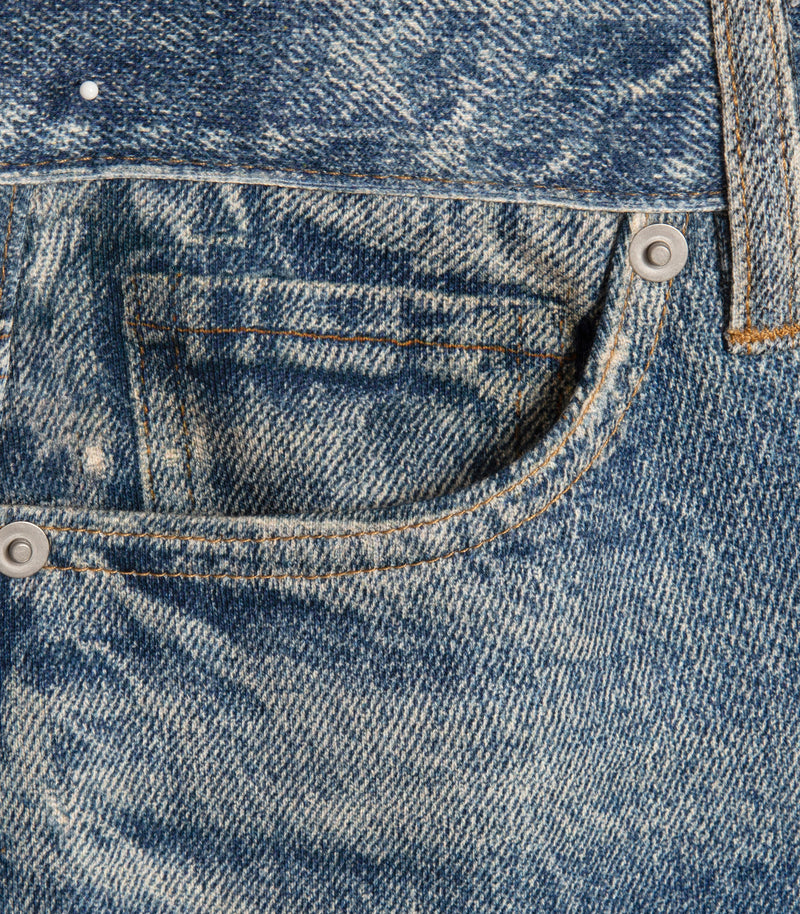 Close-up of a blue denim garment showing the front pocket and watch pocket, metal rivets, and contrast stitching.
