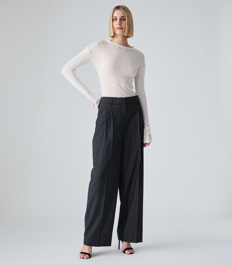 Full-length studio product view of a model presenting a sheer, lightweight white long-sleeved top. The garment features a simple crew neckline, slightly dropped shoulders, and extra-long sleeves that drape and stack softly around the hands. The semi-translucent fine-knit top is tucked into high-waisted, pleated wide-leg charcoal grey trousers and styled with minimalist black strappy stiletto sandals against a seamless light grey backdrop.