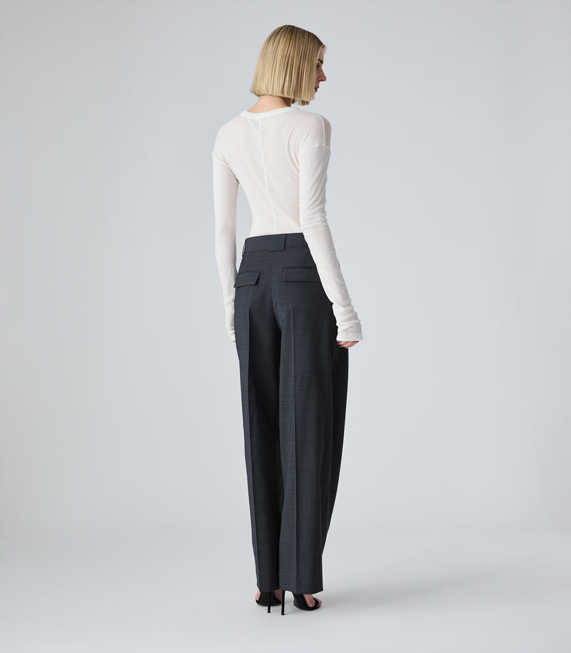 Full-length studio rear view of a model presenting a sheer, lightweight white long-sleeved top. The garment features a simple crew neckline, slightly dropped shoulders, and extra-long sleeves that drape and stack softly around the hands. The semi-translucent fine-knit top is tucked into high-waisted, pleated wide-leg charcoal grey trousers and styled with minimalist black strappy stiletto sandals against a seamless light grey backdrop.