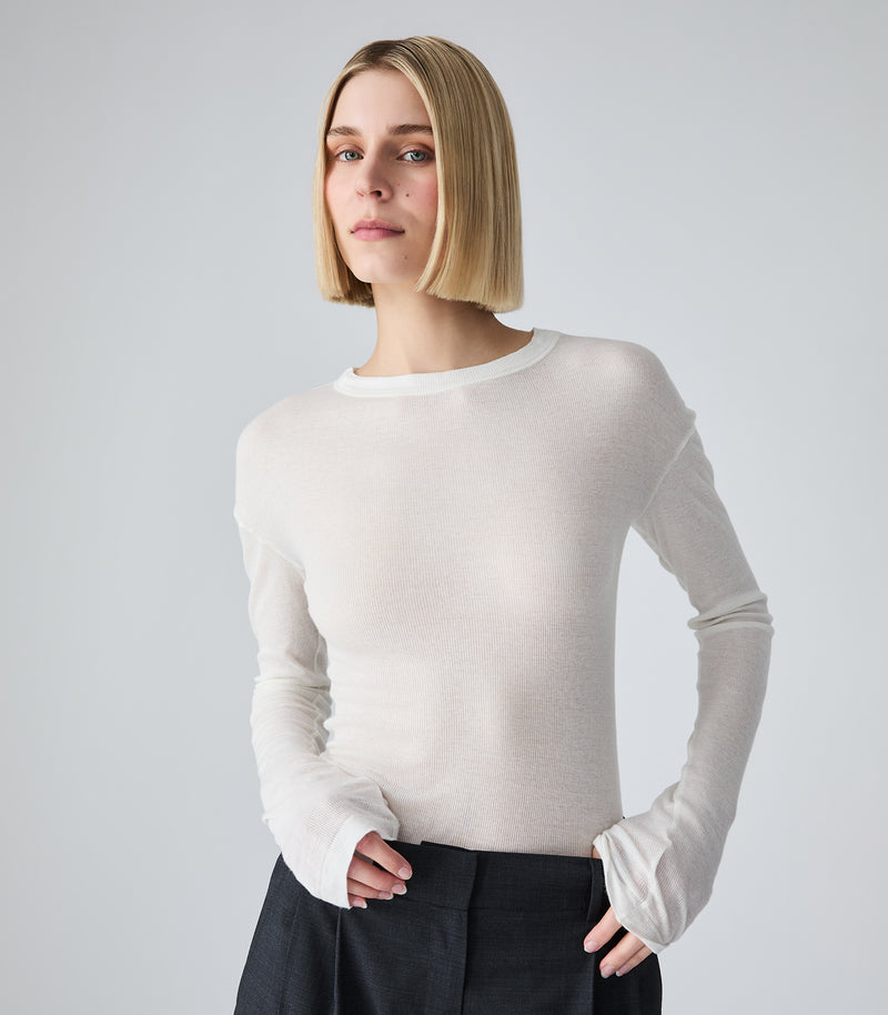 A mid-length studio product view of a model presenting a lightweight, semi-sheer white long-sleeved top. The image highlights the fine, rib-knit style texture of the fabric, a structured crew neckline with a clean banded collar, and slightly dropped shoulder seams. The extra-long cuffs drape over the hands as they rest at the waist of high-positioned charcoal grey trousers, against a plain light grey backdrop.