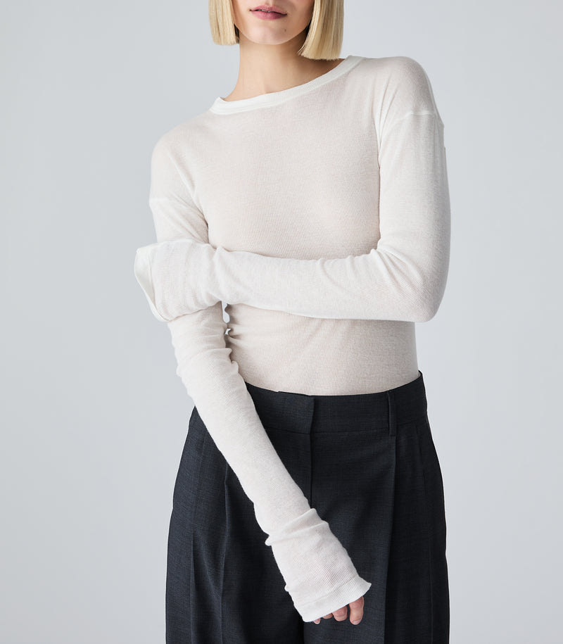 A mid-length studio product view of a model showcasing a sheer white long-sleeved crew-neck top. The model poses with her arms crossed, highlighting the lightweight, semi-translucent fine ribbed fabric and the extra-long sleeve design that gathers and stacks softly along the forearms and wrists. The garment is tucked into tailored charcoal grey wide-leg trousers, set against a seamless light grey backdrop.
