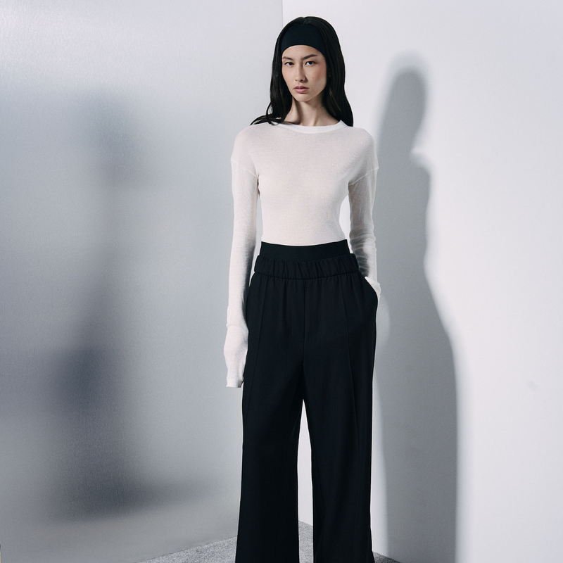 A model stands against a light gray wall wearing a fitted off-white long-sleeve top and black wide-leg trousers, with a black headband. The image has a strong shadow cast on the wall behind her.