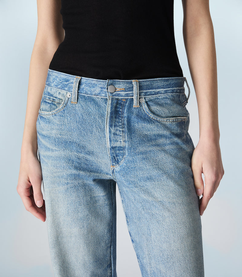 A person wearing light-wash blue denim jeans and a black sleeveless top, shown from the chest to the upper thighs against a pale blue studio background.