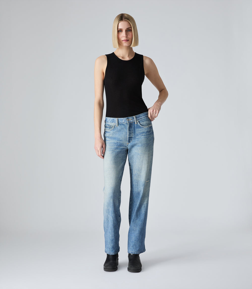 A full-length view of a model with a blunt blonde bob haircut wearing the Tessa black tank top by Vera Wang. She is styled with high-waisted, straight-leg blue jeans and black boots, posing with one hand on her hip against a solid light gray studio backdrop.