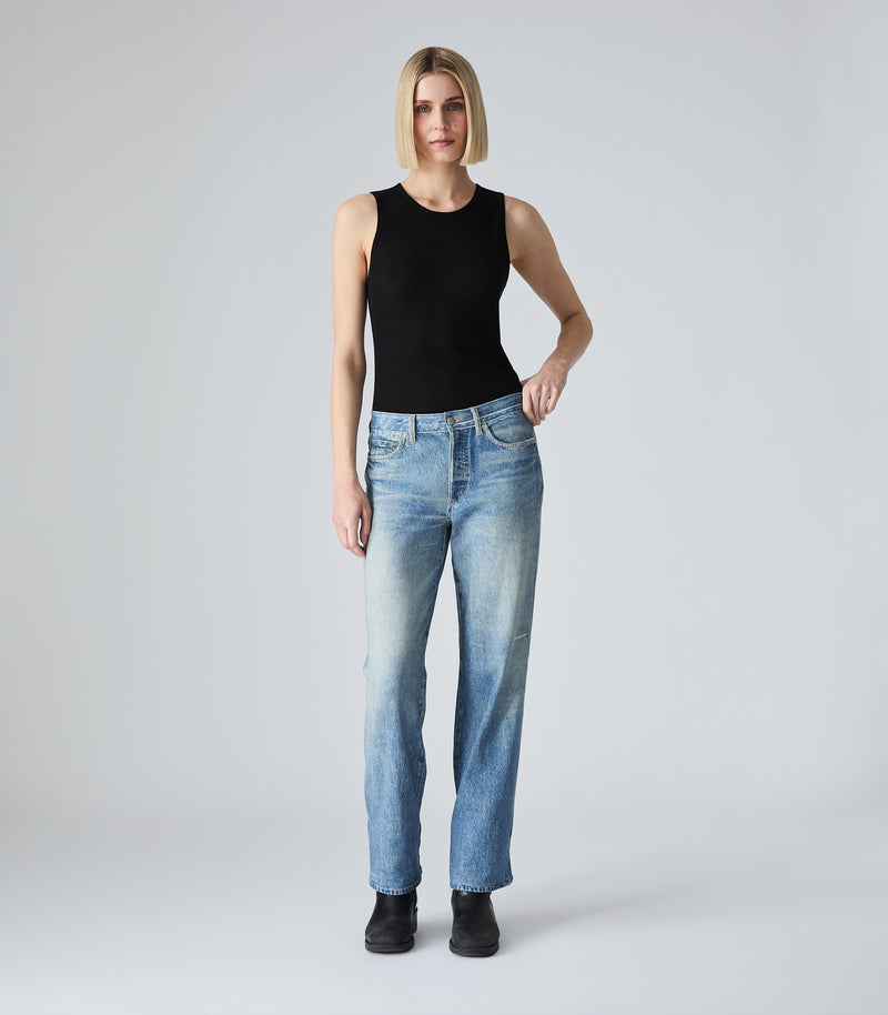 A full-length view of a model with a blunt blonde bob haircut wearing the Tessa black tank top by Vera Wang. She is styled with high-waisted, straight-leg blue jeans and black boots, posing with one hand on her hip against a solid light gray studio backdrop.