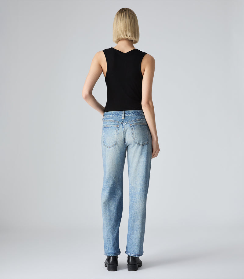 A full-length rear view of a model with a blunt blonde bob haircut, showcasing the back detail of the Tessa black tank top by Vera Wang. She is wearing mid-wash, straight-leg blue jeans and black boots, standing against a solid light gray studio backdrop.