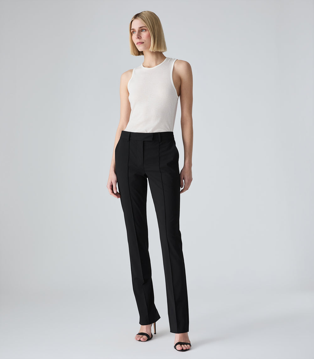 Full-length view of a model wearing a slim-fit ivory tank top featuring a classic crew neckline and a fine ribbed-knit texture. The tank top is tucked into mid-rise, slim-leg black tailored trousers designed with sharp, raised pin-tuck vertical seams along the front of each leg. The look is styled with minimalist black ankle-strap heeled sandals against a light grey background.