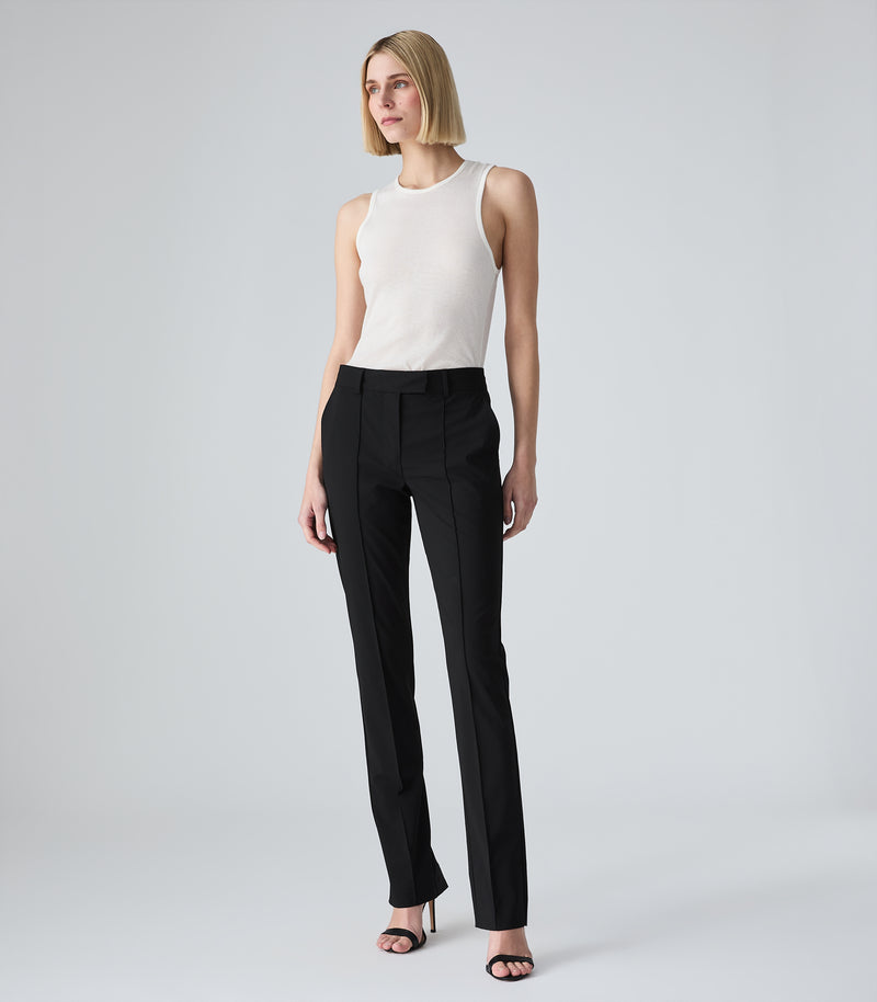 Full-length view of a model wearing a slim-fit ivory tank top featuring a classic crew neckline and a fine ribbed-knit texture. The tank top is tucked into mid-rise, slim-leg black tailored trousers designed with sharp, raised pin-tuck vertical seams along the front of each leg. The look is styled with minimalist black ankle-strap heeled sandals against a light grey background.