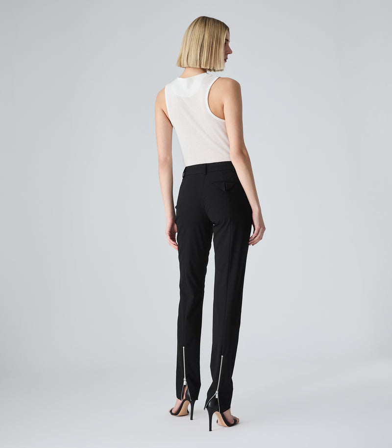 Full-length rear view of a model wearing a slim-fit ivory tank top featuring a classic racerback-inspired cut and a fine ribbed texture. The tank top is tucked into mid-rise, slim-leg black tailored trousers designed with a single asymmetric angled back flap pocket and functional silver zipper vents extending up the centre-back of each ankle hem. The look is styled with black stiletto heeled sandals against a light grey background.
