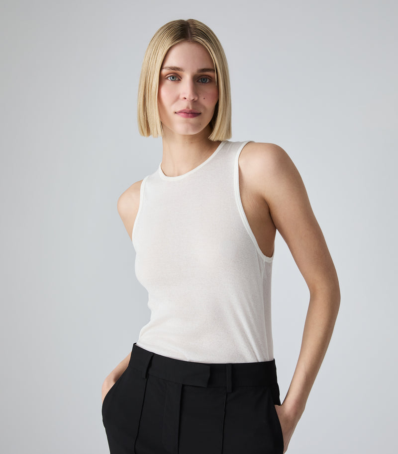 Three-quarter portrait view of a model wearing a slim-fit ivory tank top featuring a high crew neckline, athletic-style armholes, and a fine vertical ribbed-knit texture. The model is standing with her hands in the pockets of mid-rise black tailored trousers, showcasing the close fit of the top against a light grey background.