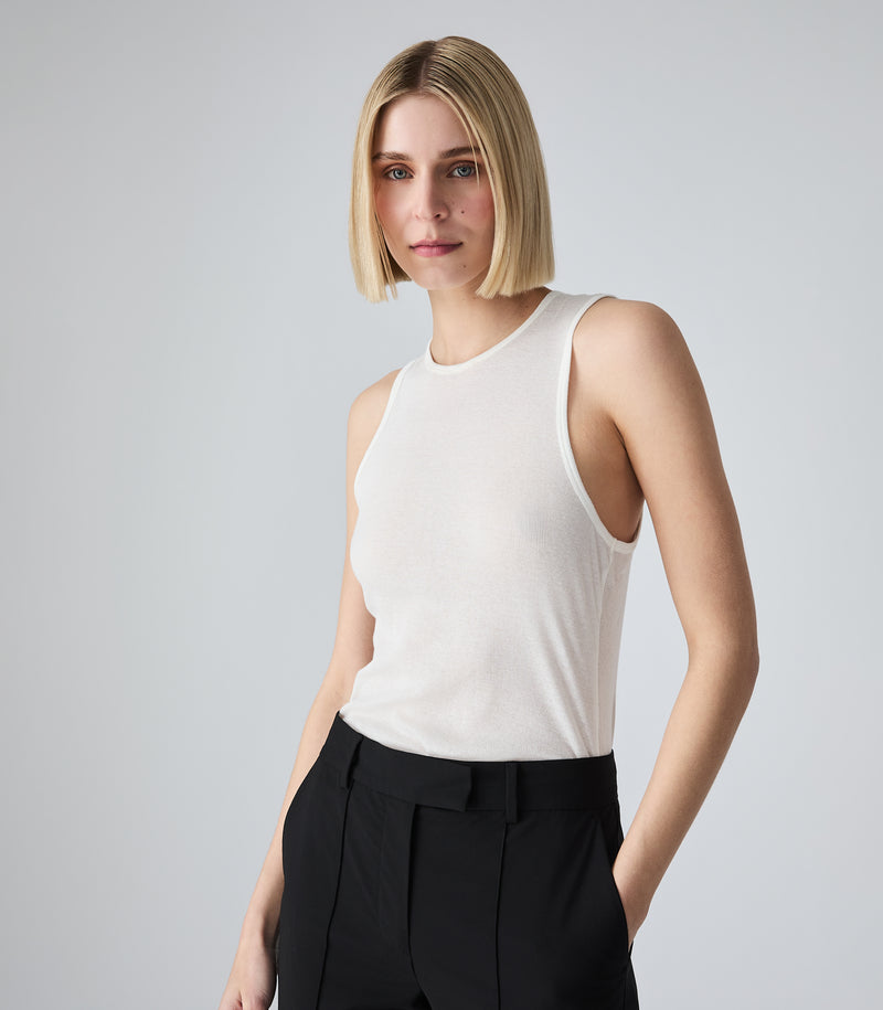Three-quarter portrait view of a model wearing a slim-fit ivory tank top featuring a high crew neckline, athletic-style armholes, and a fine vertical ribbed-knit texture. The model is standing with her hands in the pockets of mid-rise black tailored trousers, showcasing the close fit of the top against a light grey background.
