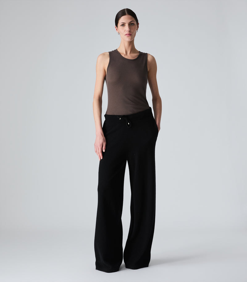 A model stands against a light gray studio background wearing a fitted sleeveless brown top and black wide-leg trousers.