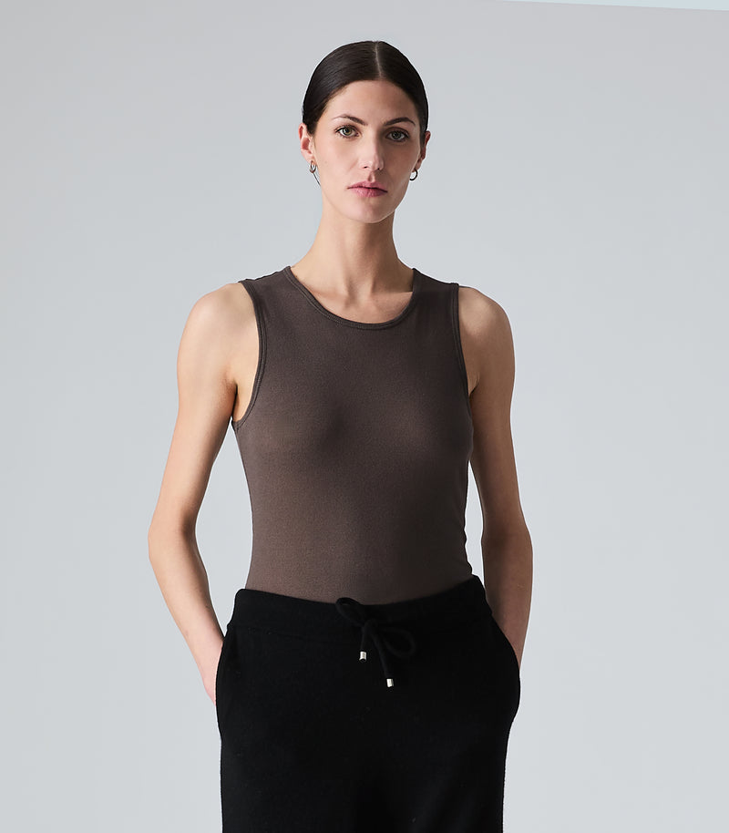 A woman modeling a sleeveless, crew-neck tank top in a dark brown color, paired with black drawstring pants, against a solid light gray background.