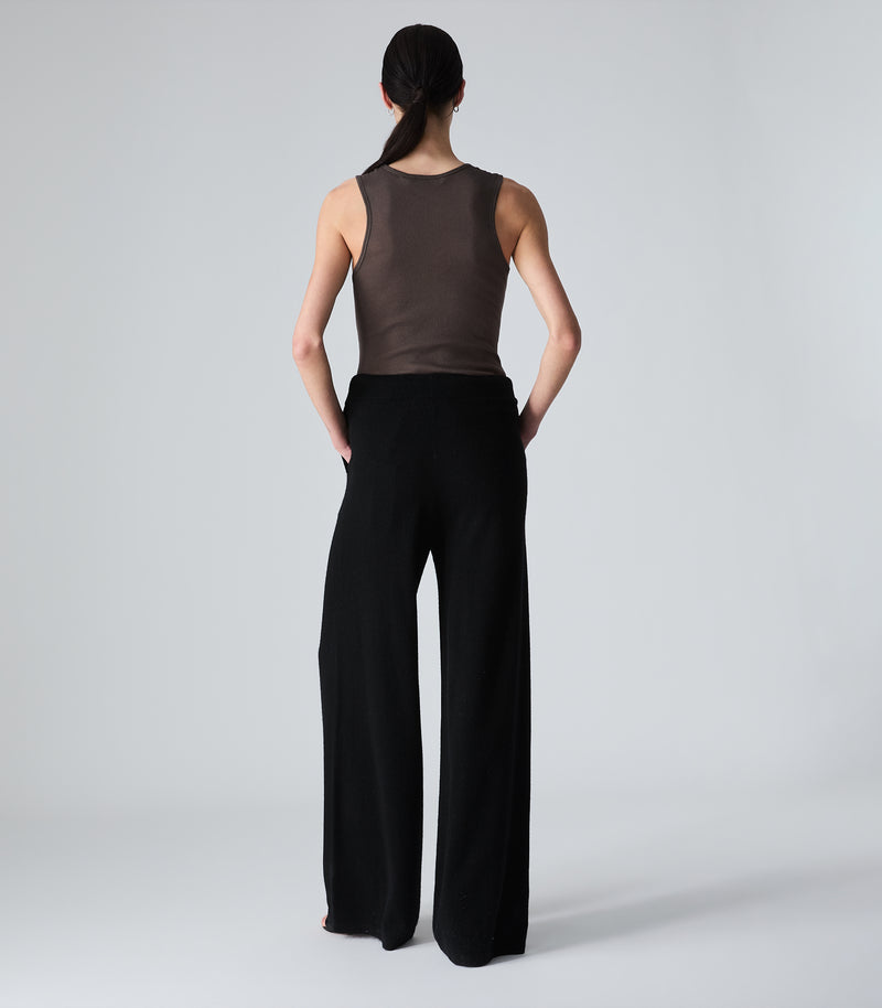 A model shown from the back wearing a sleeveless brown mesh top and black wide-leg pants, standing against a light gray studio background.