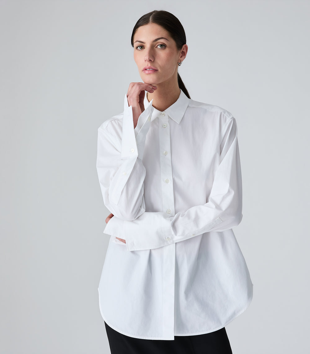 A woman modeling a white long-sleeve button-up shirt with a relaxed, slightly oversized fit, paired with black pants, against a light gray studio background.