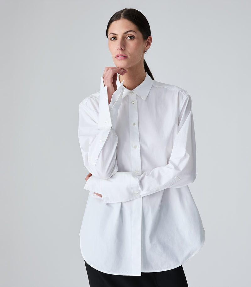 A woman modeling a white long-sleeve button-up shirt with a relaxed, slightly oversized fit, paired with black pants, against a light gray studio background.
