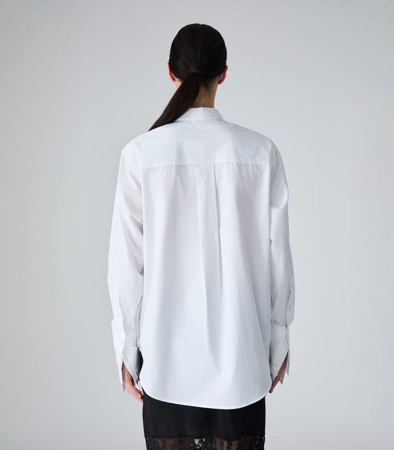 A woman viewed from the back wearing a white long-sleeve button-up shirt and a black skirt, set against a plain light gray studio background.