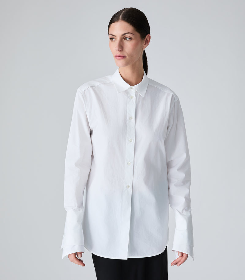 A woman modeling a white long-sleeve button-up shirt with a pointed collar, photographed against a plain light gray studio background.