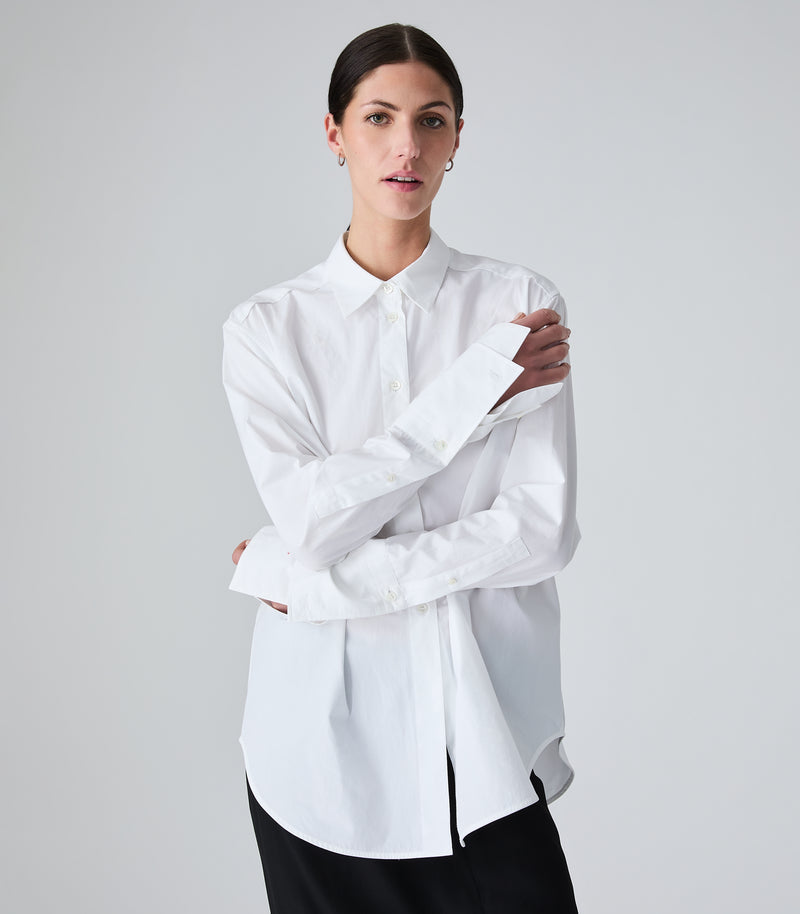 A woman modeling a white long-sleeve button-up shirt with a relaxed, slightly oversized fit and a subtle side slit, paired with black bottoms, against a light gray studio background.