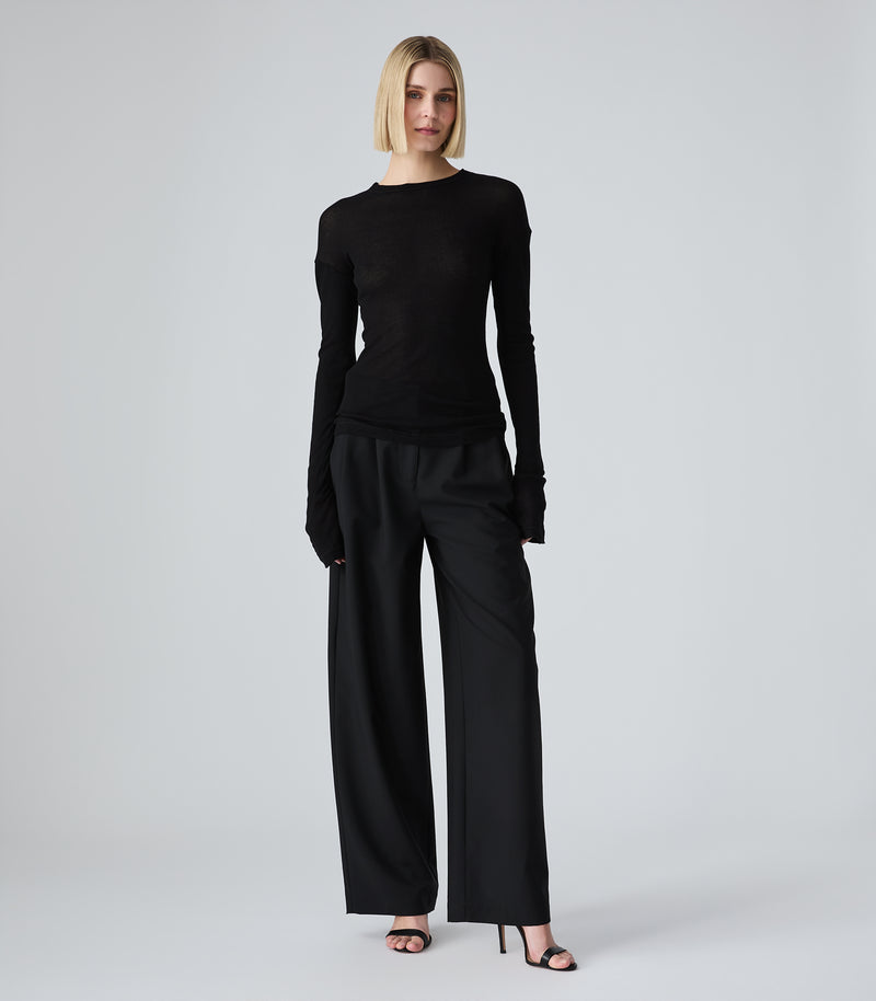 A model stands against a light gray studio background wearing a fitted black long-sleeve top and black wide-leg trousers with black high heels.