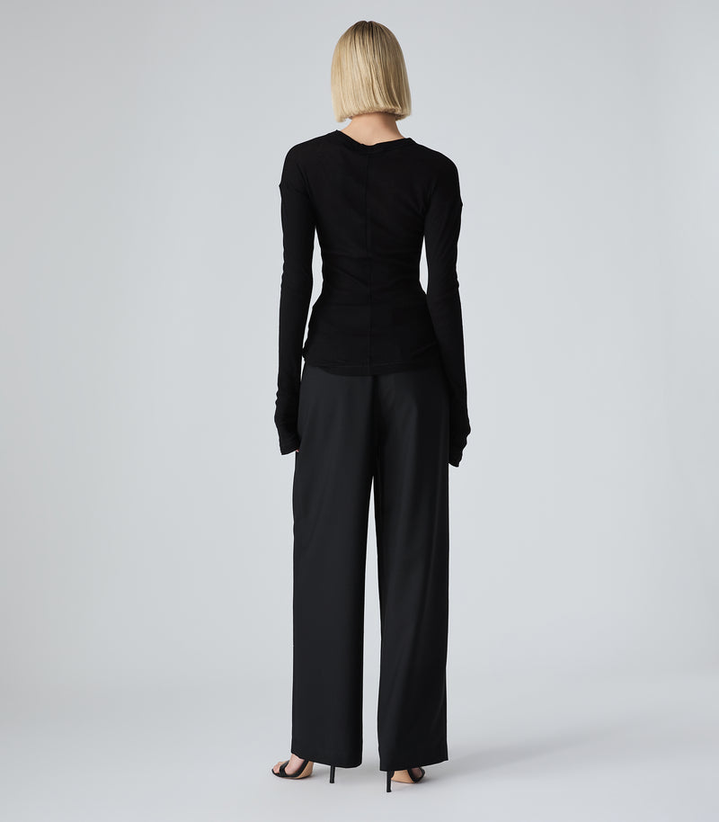 A rear view of a model standing against a light gray studio background wearing a fitted black long-sleeve top and black wide-leg trousers with black high heels.