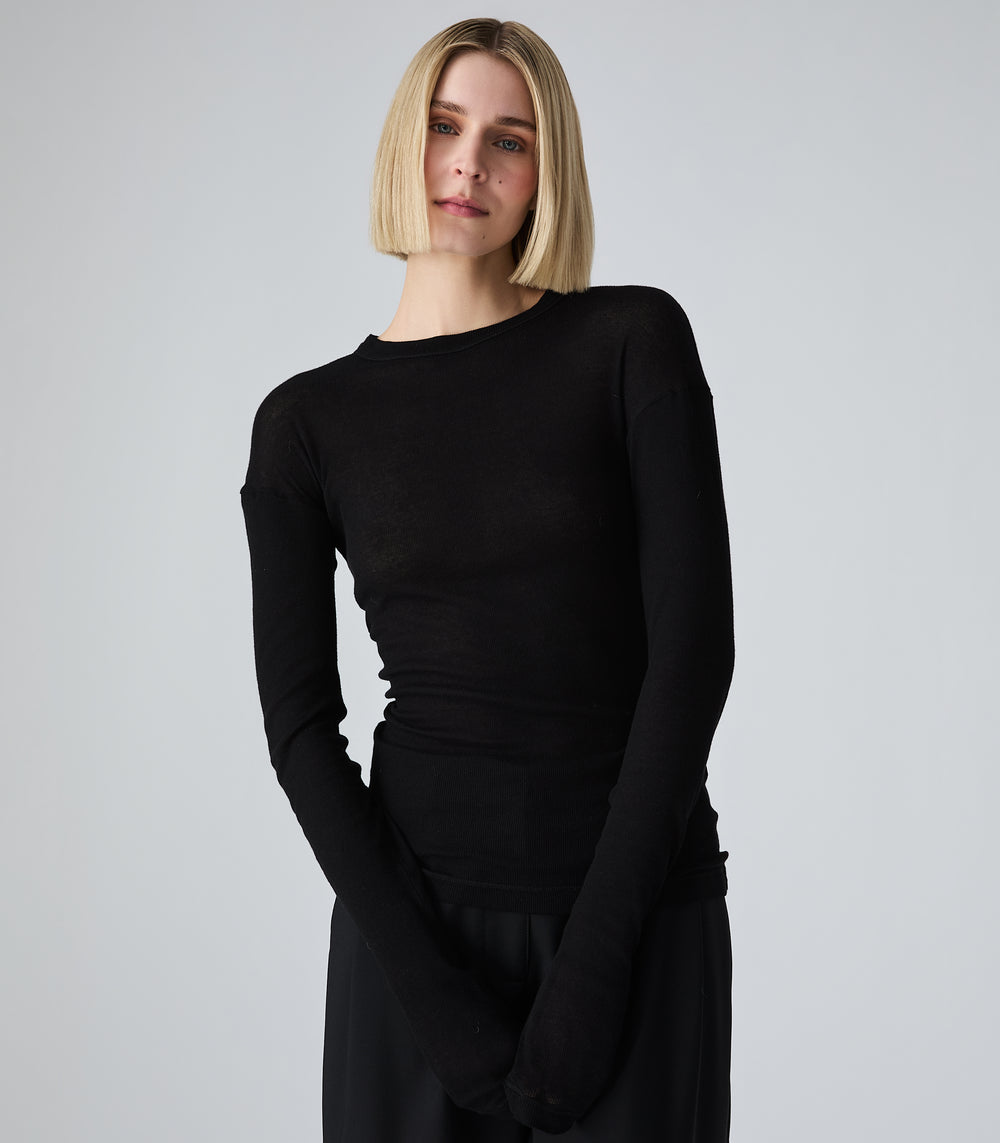 A woman with a short blonde bob wearing a fitted black long-sleeve top and black skirt, standing against a light gray studio background.