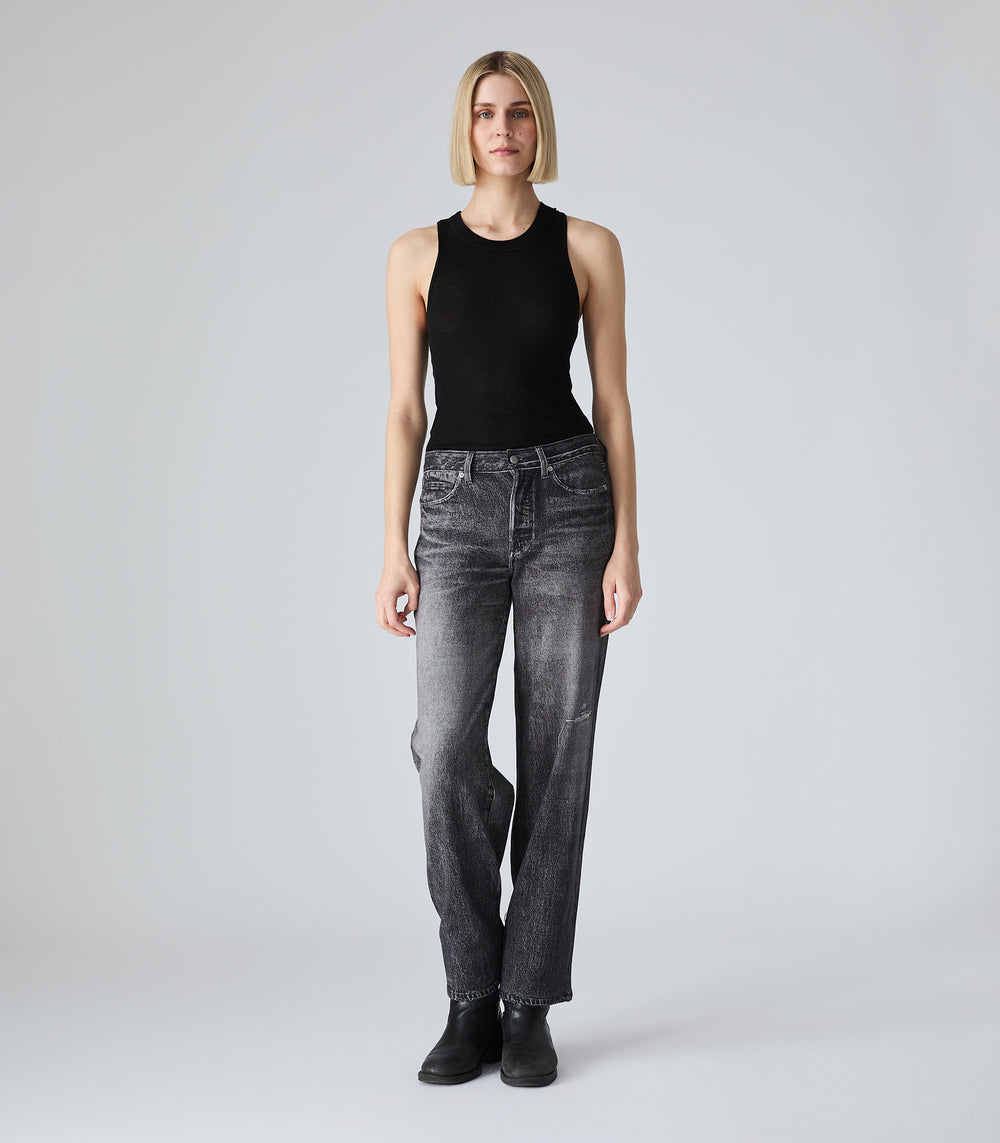 Full-length view of a model wearing a slim-fit black tank top featuring a classic crew neckline and a lightweight, slightly sheer fine ribbed-knit texture. The tank top is tucked into mid-rise, straight-leg Grey jeans. The tonal look is styled with minimalist black heeled boots against a light grey background.