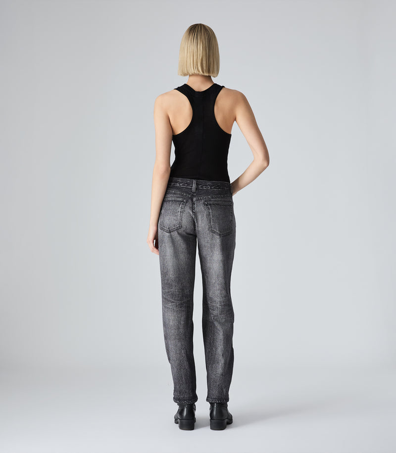 Full-length rear view of a model wearing a slim-fit black tank top featuring a racerback silhouette and a lightweight, semi-sheer fine ribbed-knit texture. The tank top is tucked into mid-rise, slim-leg black tailored trousers. The monochromatic look is styled with black heeled boots against a light grey background.