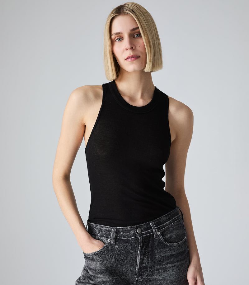 A woman with a blunt, straight blonde bob hairstyle wearing a ribbed black racerback tank top. She is posing against a solid light gray background with one hand tucked into the pocket of her dark wash, acid-washed denim jeans.