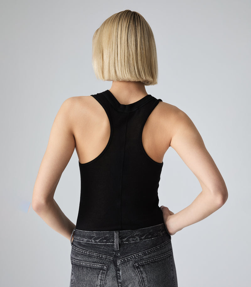 Rear view of a woman with a straight, blunt blonde bob haircut, showing the back detail of a black ribbed racerback tank top with a subtle center seam. She has her hands on her hips, wearing dark wash, acid-washed denim jeans against a solid light gray background.