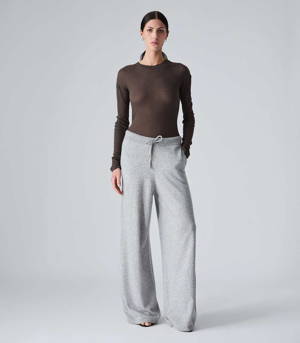 A woman modeling a dark brown long-sleeve mock neck top paired with light gray wide-leg sweatpants, standing against a neutral light gray studio background.