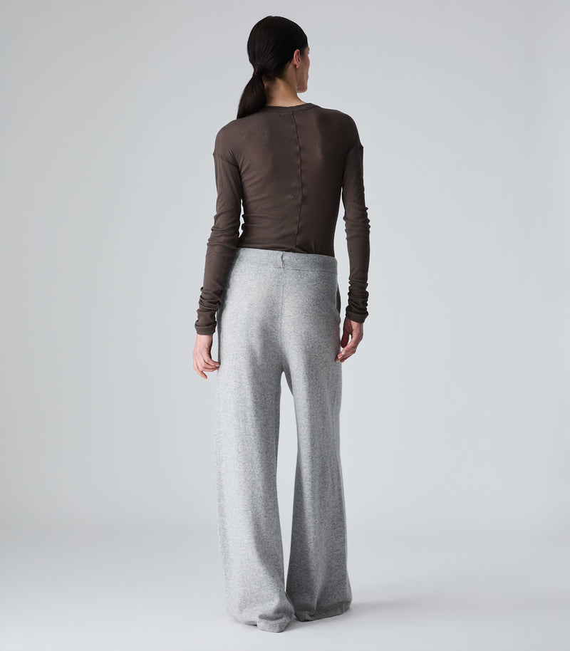 A woman shown from the back wearing a dark brown long-sleeve top and light gray wide-leg pants against a plain light gray studio background.