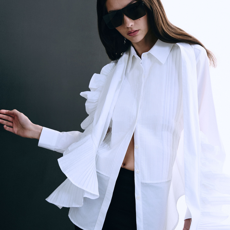 A woman wearing a white long-sleeve pleated shirt with dramatic ruffle details on the sleeves and collar, styled with black sunglasses and black bottoms against a dark gray background.