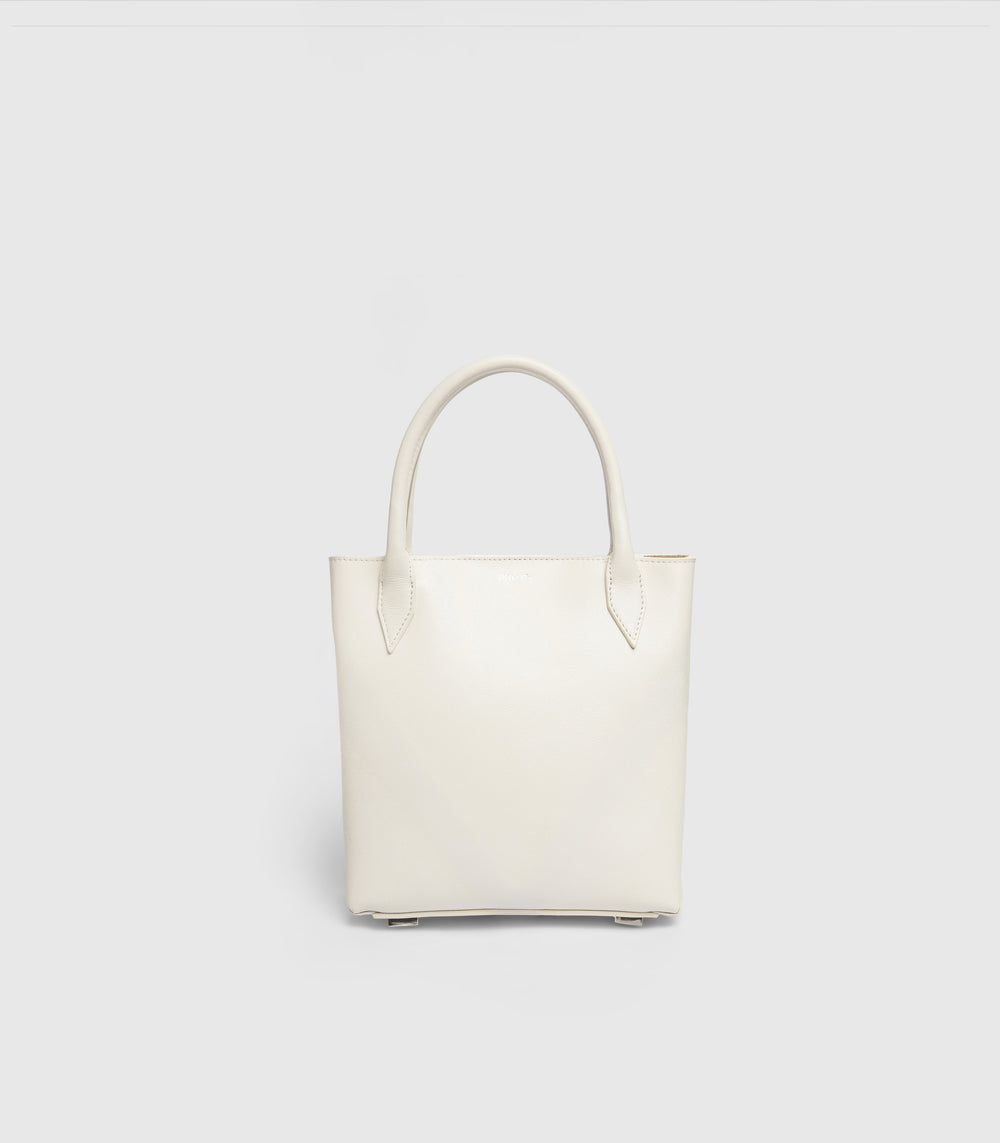 A minimalist off-white leather tote bag with double top handles, shown against a solid light gray background.