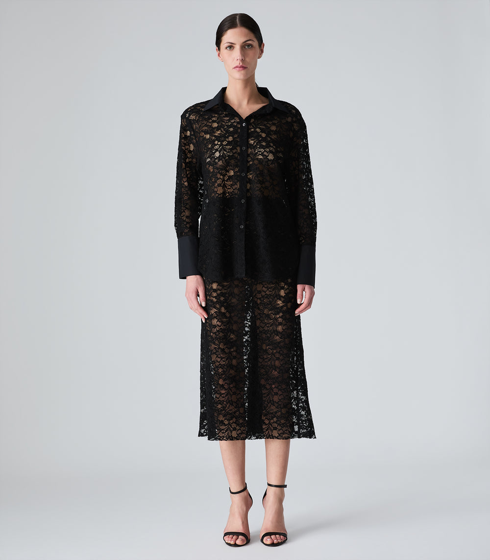 A model stands against a light gray studio background wearing a black sheer lace long-sleeve top and matching midi skirt with solid black trim.