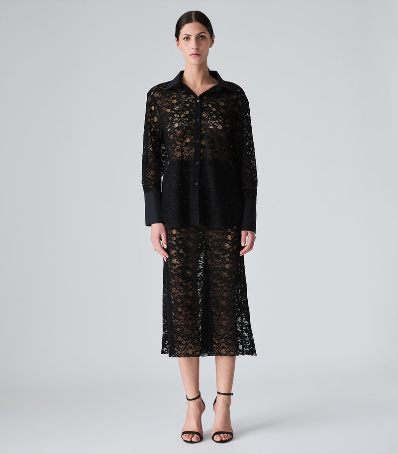 A model stands against a light gray studio background wearing a black sheer lace long-sleeve top and matching midi skirt with solid black trim.