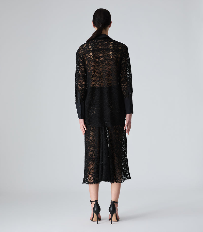 A model viewed from the back wearing a black lace long-sleeve jacket and matching midi skirt, paired with black high-heeled sandals, against a light gray studio background.