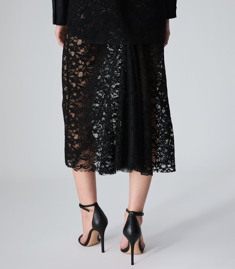 A person shown from the back wearing a black lace midi skirt with a sheer floral pattern, paired with black high-heeled sandals, against a light gray studio background.