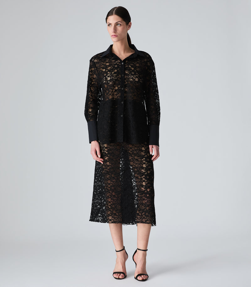 A model stands against a light gray studio background wearing a black sheer lace long-sleeve shirt and matching midi skirt with solid black underlayers and black strappy heels.