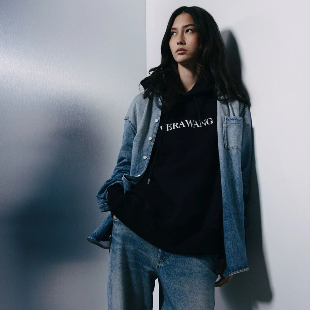 A model wearing a black hoodie with 'VERAWANG' printed in white across the chest, layered under a light blue denim jacket and paired with light-wash jeans, leaning against a gray wall.