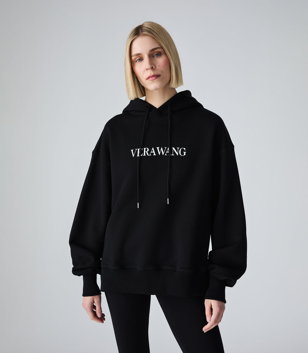 A closer-shot of a female model wearing a black oversized hoodie with a white 'VERA WANG' spell-out logo across the chest, paired with black leggings, against a white background.