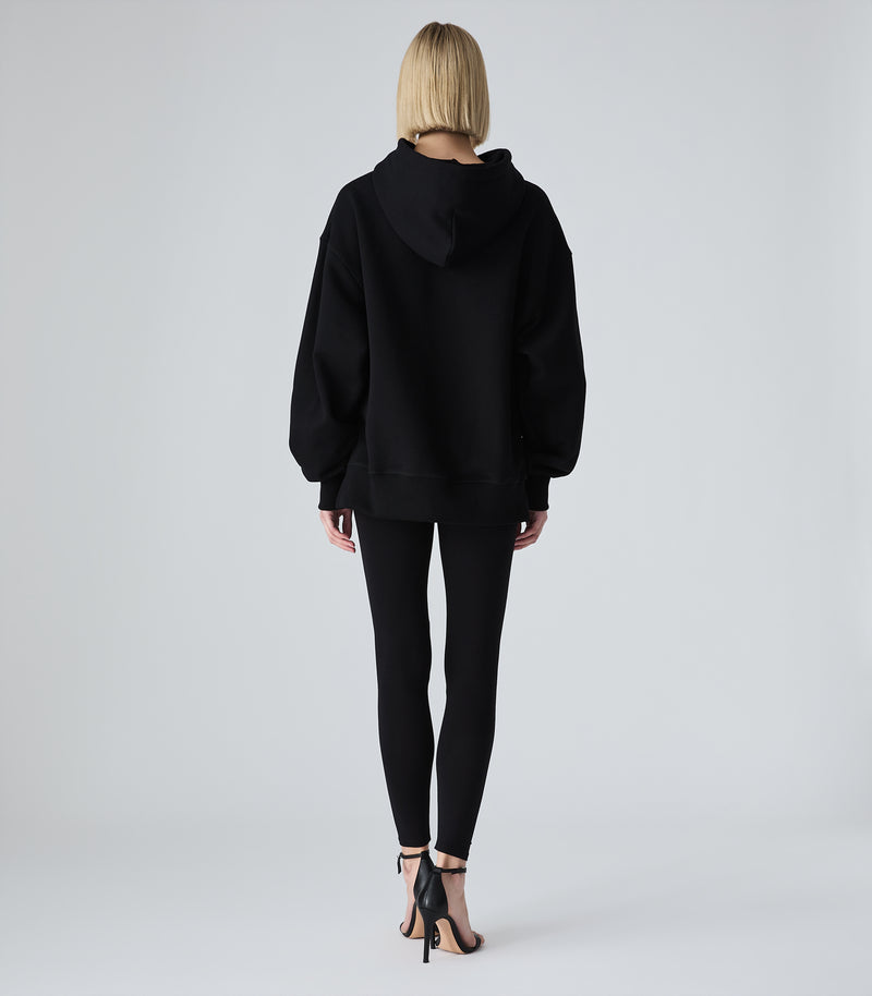 Female model wearing a black oversized hoodie, paired with black leggings and heels, against a white background.