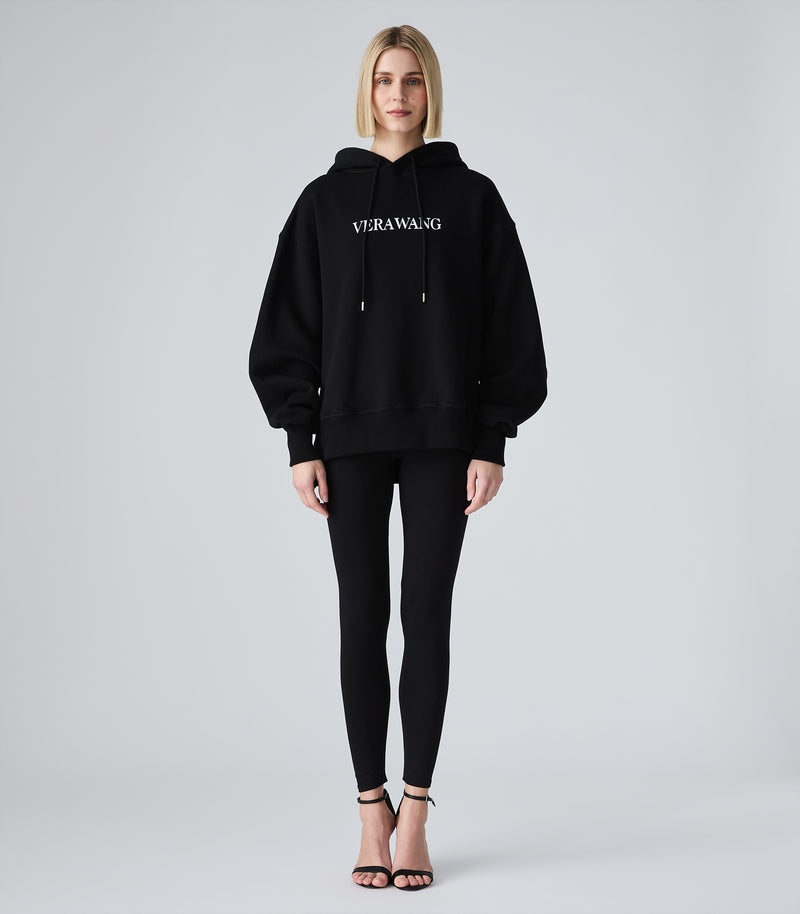 Female model wearing a black oversized hoodie with a white 'VERA WANG' spell-out logo across the chest, paired with black leggings and heels, against a white background.