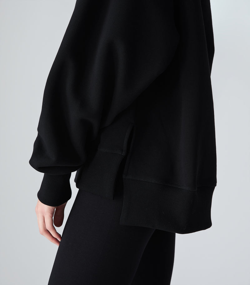 Close-up side view of a person wearing an oversized black sweatshirt with ribbed cuffs and a side-slit hem, paired with black leggings against a white background.