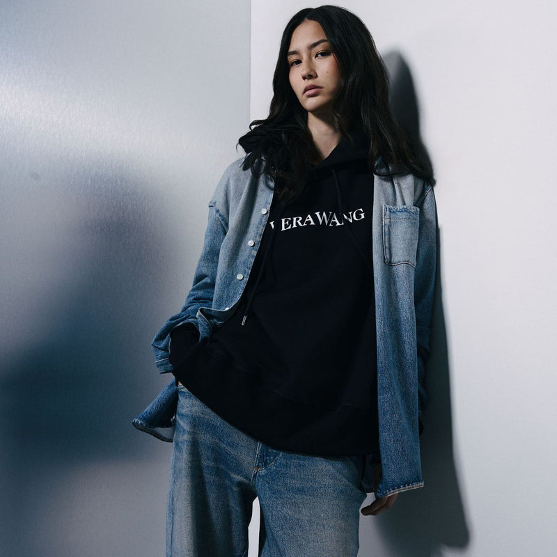 A woman leans against a white wall wearing a black hoodie with 'VERAWANG' printed in white across the chest, layered under an open light-wash denim jacket and paired with light-wash jeans.