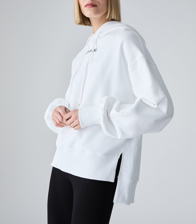 A model wearing a white oversized pullover hoodie with a small black text graphic on the left chest, paired with black leggings, against a light gray studio background.