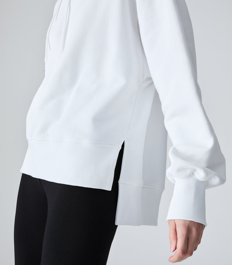 A close-up of a person wearing a white long-sleeve sweatshirt with black leggings, showing the side profile and ribbed cuffs against a light gray studio background.