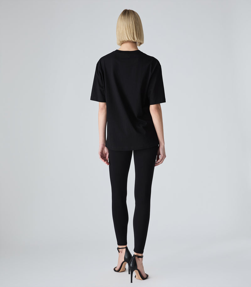 Full-length back view of a model wearing a loose-fit black crewneck t-shirt. The t-shirt is styled with black leggings and black strappy stiletto heels, against a white background.