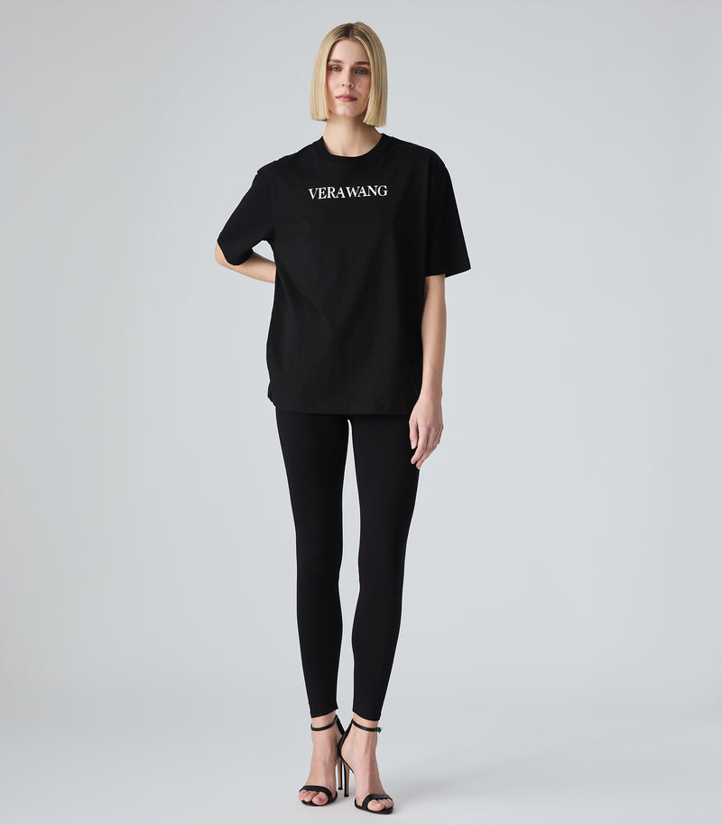 Full-length view of a model wearing a loose-fit black crewneck t-shirt featuring 'VERA WANG' spell-out logo in white lettering across the chest. The t-shirt is styled with black leggings and black strappy stiletto heels, against a white background.
