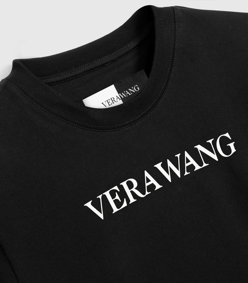 Close-up, angled product view of a black cotton crewneck t-shirt, highlighting the white printed 'VERA WANG' logo across the upper chest, the ribbed knit collar, and a woven black-and-white inner neck label.